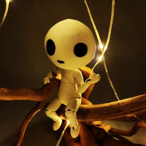 May include: A white, articulated doll with large black eyes and a small mouth sits on a brown branch. The doll is illuminated by small, warm-toned fairy lights, creating a whimsical and enchanting scene. The doll's pose suggests a sense of curiosity.