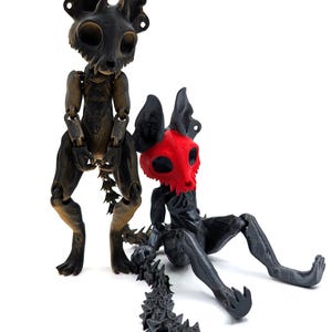 May include: Two articulated black and gold skeletal figures with long tails. The figure on the left is standing and the figure on the right is sitting with its tail extended behind it. The figure on the right has a red skull face.