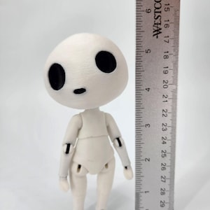 May include: A small, articulated doll with a rounded head and large black eyes. The doll is off-white and stands approximately 5 inches tall, measured with a ruler. The doll's limbs are jointed, allowing for posing. The ruler is marked in inches.