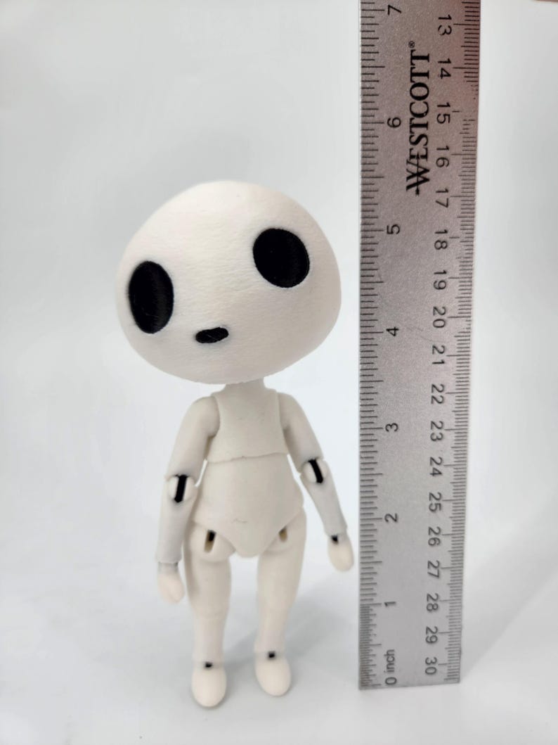 May include: A small, articulated doll with a rounded head and large black eyes. The doll is off-white and stands approximately 5 inches tall, measured with a ruler. The doll's limbs are jointed, allowing for posing. The ruler is marked in inches.