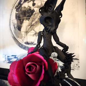 May include: A black articulated doll with a skull-like head and large ears sits on a rose. The doll is posed with one leg extended and the other bent. The doll is sitting on a rose with a black and white background.