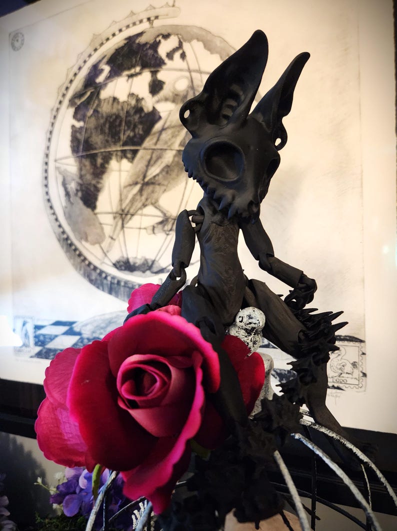 May include: A black articulated doll with a skull-like head and large ears sits on a rose. The doll is posed with one leg extended and the other bent. The doll is sitting on a rose with a black and white background.
