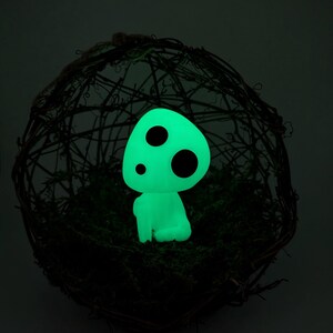 K&ouml;nnte beinhalten: Eine leuchtend gr&uuml;ne Figur mit drei schwarzen Punkten, die einem Waldgeist &auml;hnelt, sitzt auf gr&uuml;nem Moos in einer gewebten, kugelf&ouml;rmigen Struktur. Die Figur strahlt sanft vor einem dunklen Hintergrund.