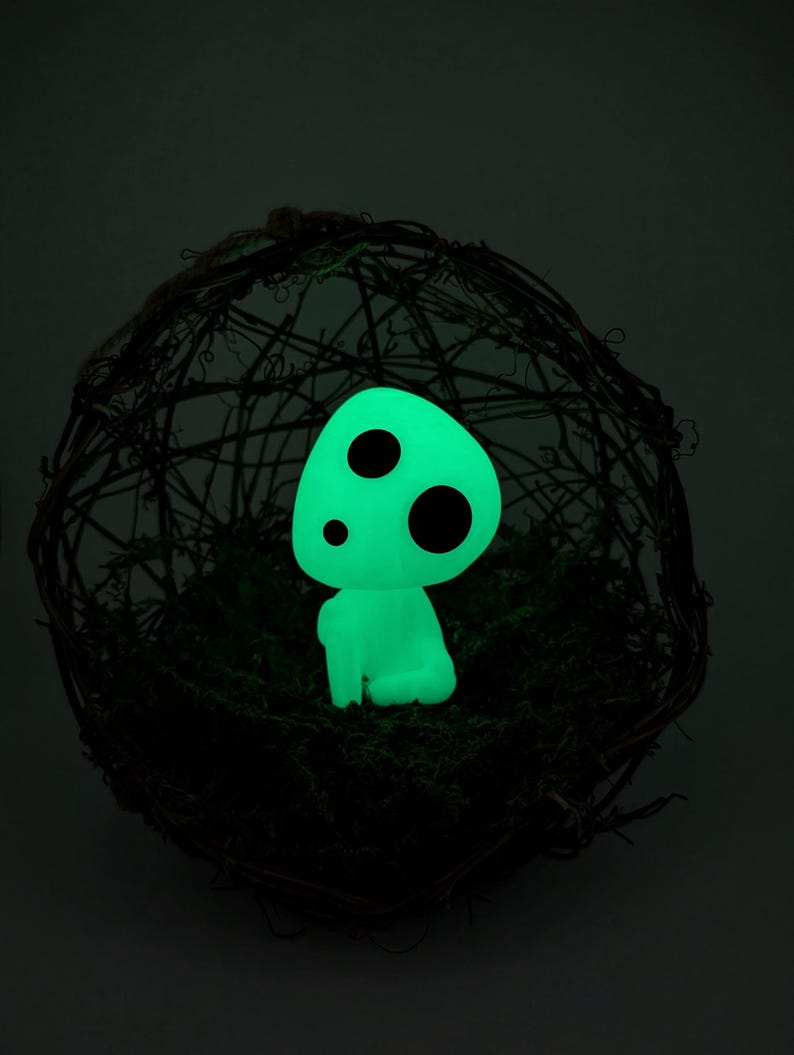 K&ouml;nnte beinhalten: Eine leuchtend gr&uuml;ne Figur mit drei schwarzen Punkten, die einem Waldgeist &auml;hnelt, sitzt auf gr&uuml;nem Moos in einer gewebten, kugelf&ouml;rmigen Struktur. Die Figur strahlt sanft vor einem dunklen Hintergrund.