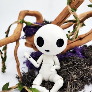 May include: A white doll with a round head and large black eyes, posed in a seated position. The doll is surrounded by natural elements, including brown branches, purple moss, and green vines. The doll's arms and legs are articulated.