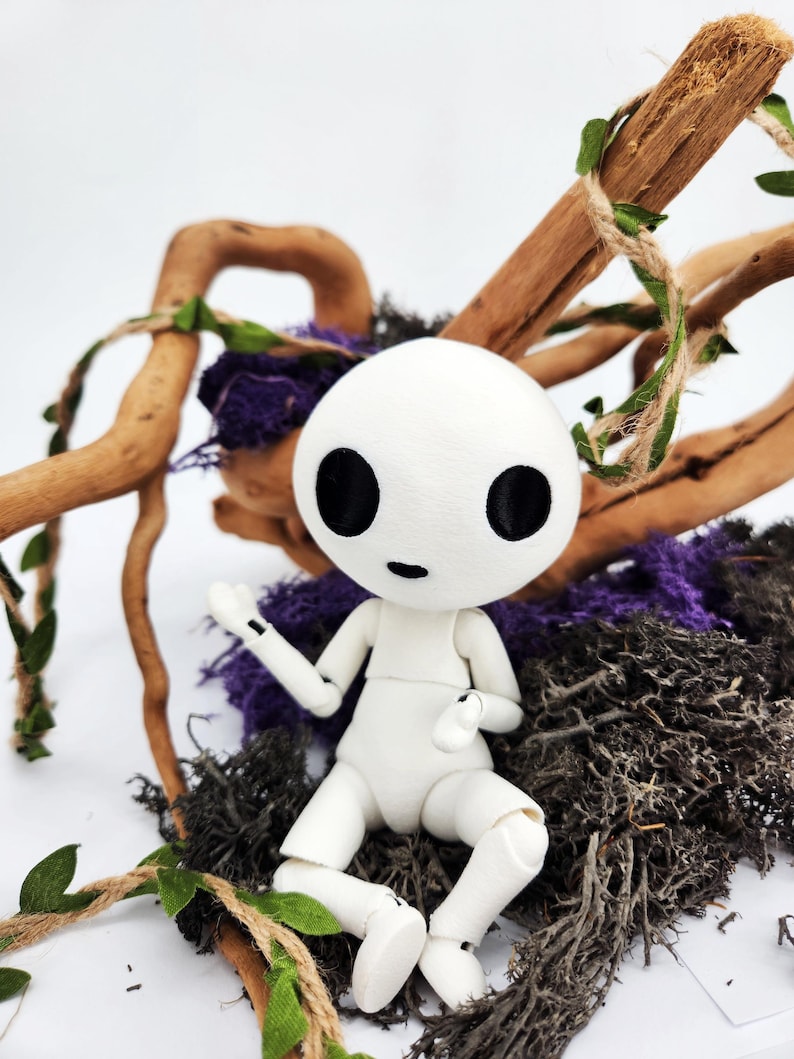 May include: A white doll with a round head and large black eyes, posed in a seated position. The doll is surrounded by natural elements, including brown branches, purple moss, and green vines. The doll's arms and legs are articulated.