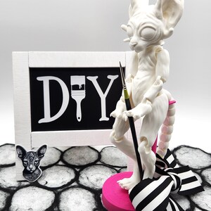 May include: Unpainted, articulated doll of a hairless cat, standing on a pink base with a black and white ribbon. The doll is holding a paintbrush and is standing in front of a white frame with a black chalkboard that says "DIY" with a paintbrush icon.