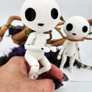 May include: Two white articulated dolls with large black eyes and small mouths. One doll is posed in a sitting position, while the other stands nearby. The dolls are set against a backdrop of branches and purple foliage, creating a whimsical scene.