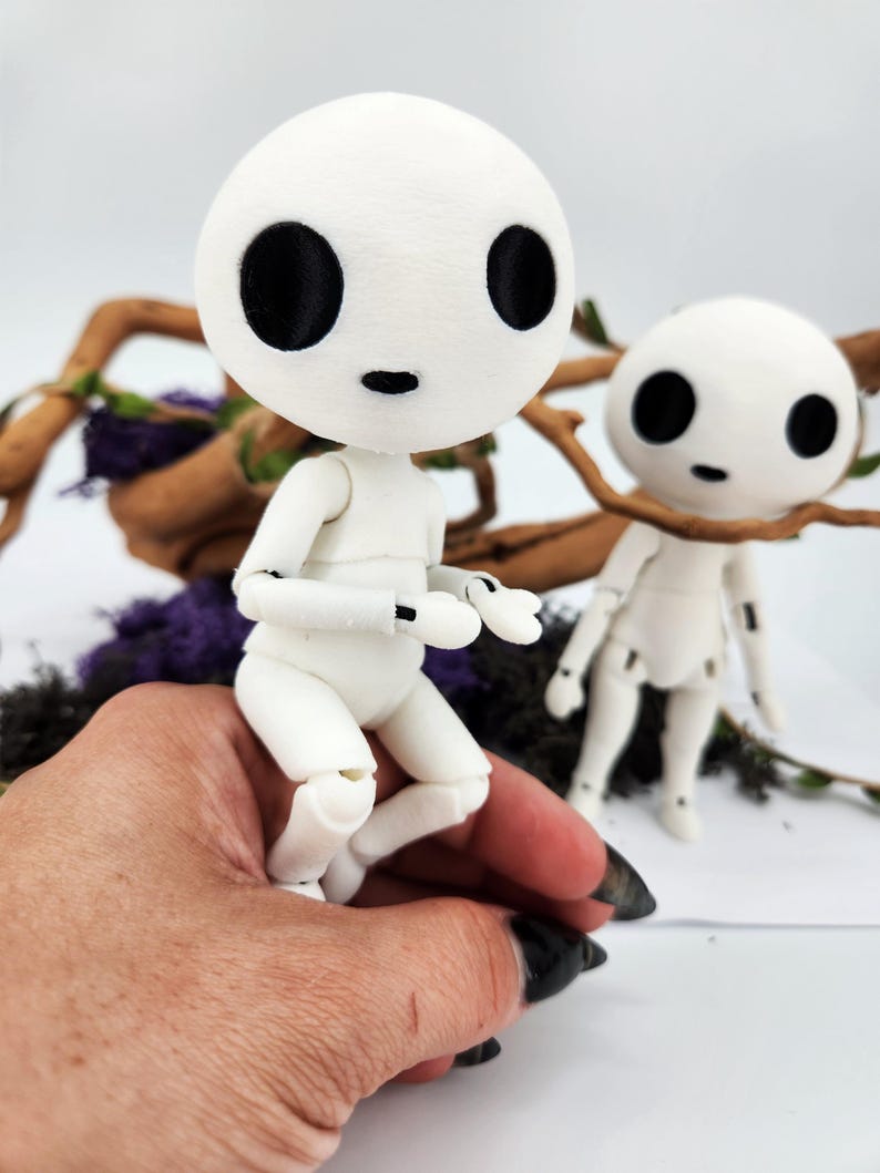 May include: Two white articulated dolls with large black eyes and small mouths. One doll is posed in a sitting position, while the other stands nearby. The dolls are set against a backdrop of branches and purple foliage, creating a whimsical scene.