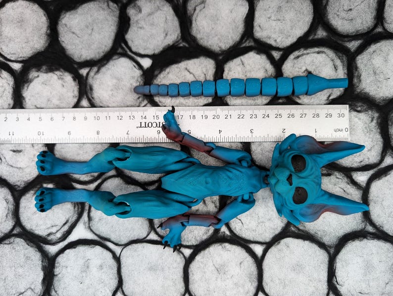 May include: A blue articulated doll with a long tail, a cat-like face, and large ears. The doll is lying on a black and white textured surface.