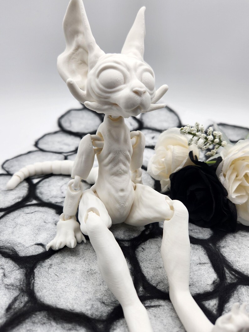 May include: A white, articulated, doll with a cat-like face and a long tail. The doll is sitting on a black and white patterned surface.