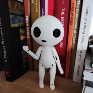 May include: A white articulated figure with a round head and large black eyes stands on a wooden surface. The figure has jointed arms and legs, and is posed with one arm raised. Books of various colors and titles are in the background.