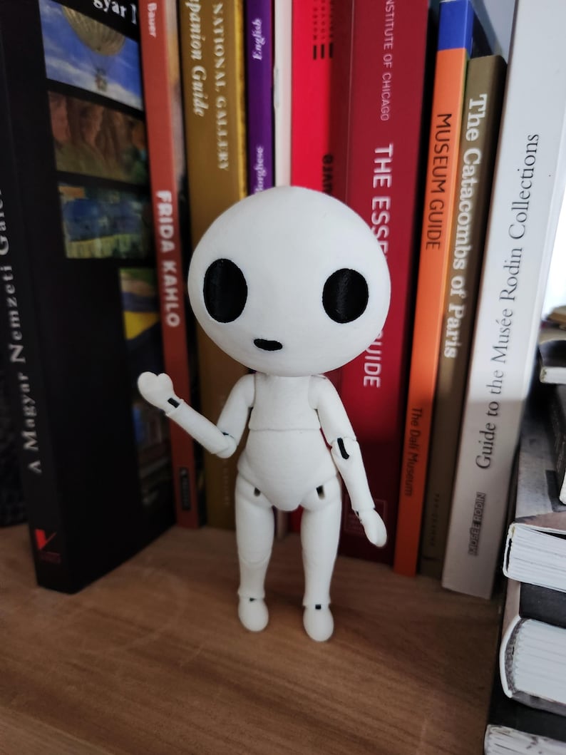 May include: A white articulated figure with a round head and large black eyes stands on a wooden surface. The figure has jointed arms and legs, and is posed with one arm raised. Books of various colors and titles are in the background.