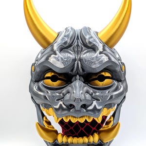 May include: A gray and gold Oni mask with sharp teeth and horns. The mask has a detailed design and is made of a durable material.