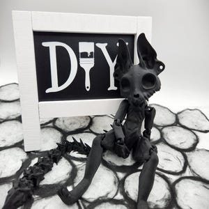 May include: A black skeletal figure with a long, spiky tail sits on a black and white patterned surface. A white sign with the word "DIY" and a paintbrush icon is behind the figure.