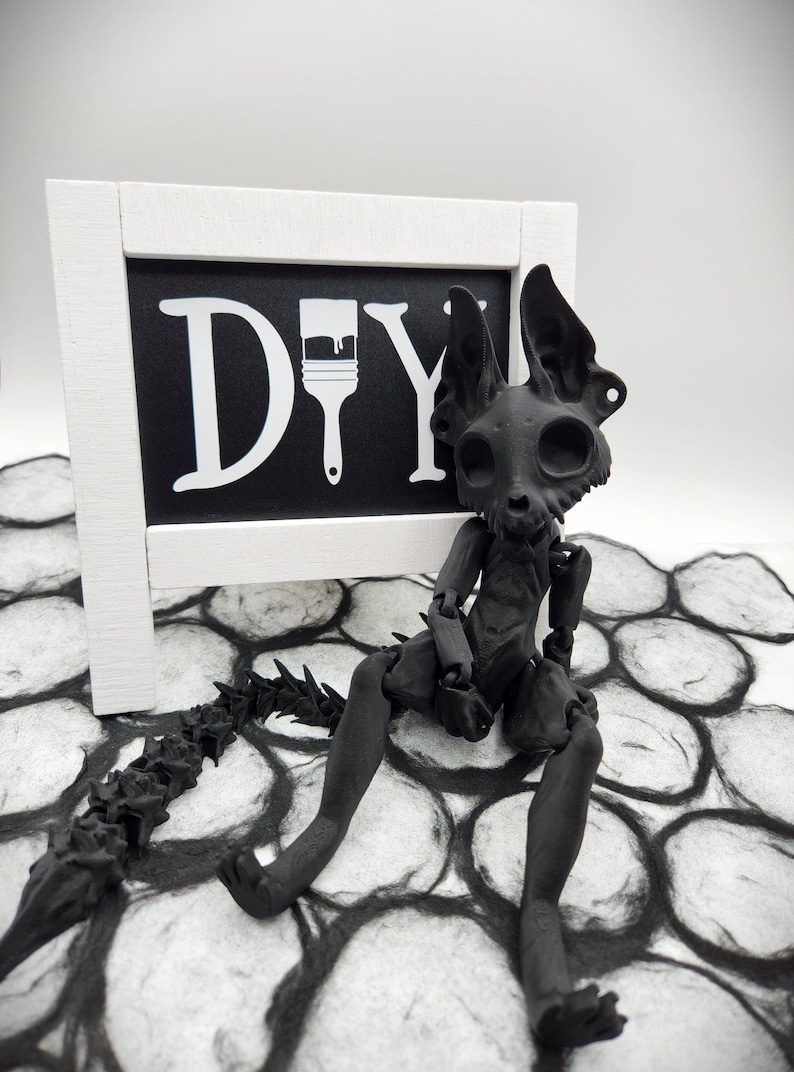 May include: A black skeletal figure with a long, spiky tail sits on a black and white patterned surface. A white sign with the word "DIY" and a paintbrush icon is behind the figure.