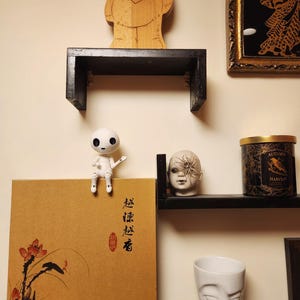 May include: A display of decorative items on black shelves against a beige wall. Items include a wooden doll, a small white doll, a box with Asian-style art, a candle, a ceramic head, and a white face-shaped cup.
