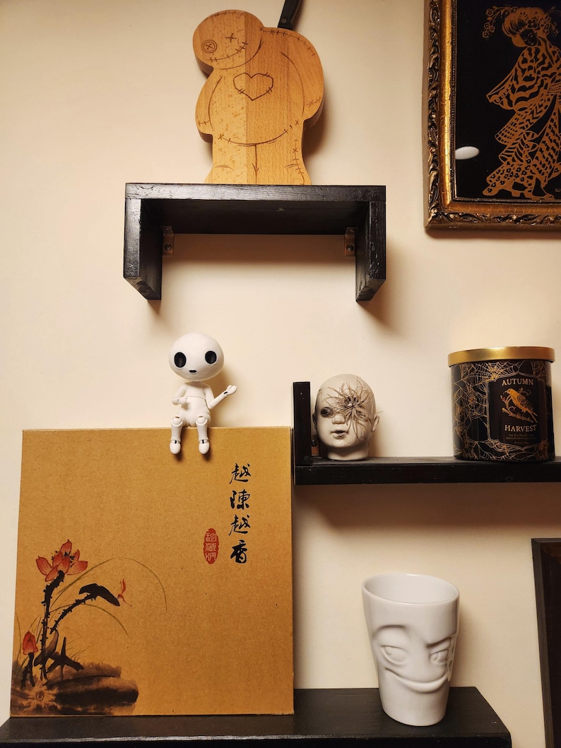 May include: A display of decorative items on black shelves against a beige wall. Items include a wooden doll, a small white doll, a box with Asian-style art, a candle, a ceramic head, and a white face-shaped cup.