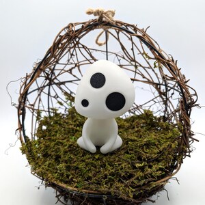 K&ouml;nnte beinhalten: Eine wei&szlig;e Figur mit drei schwarzen Punkten, die an einen Waldgeist erinnert, sitzt auf einem Bett aus gr&uuml;nem Moos in einer gewebten Zweigkugel. Die Kugel wird von einer nat&uuml;rlichen Schnurschlaufe gehalten, was eine skurrile, von der Natur inspirierte Darstellung ergibt.