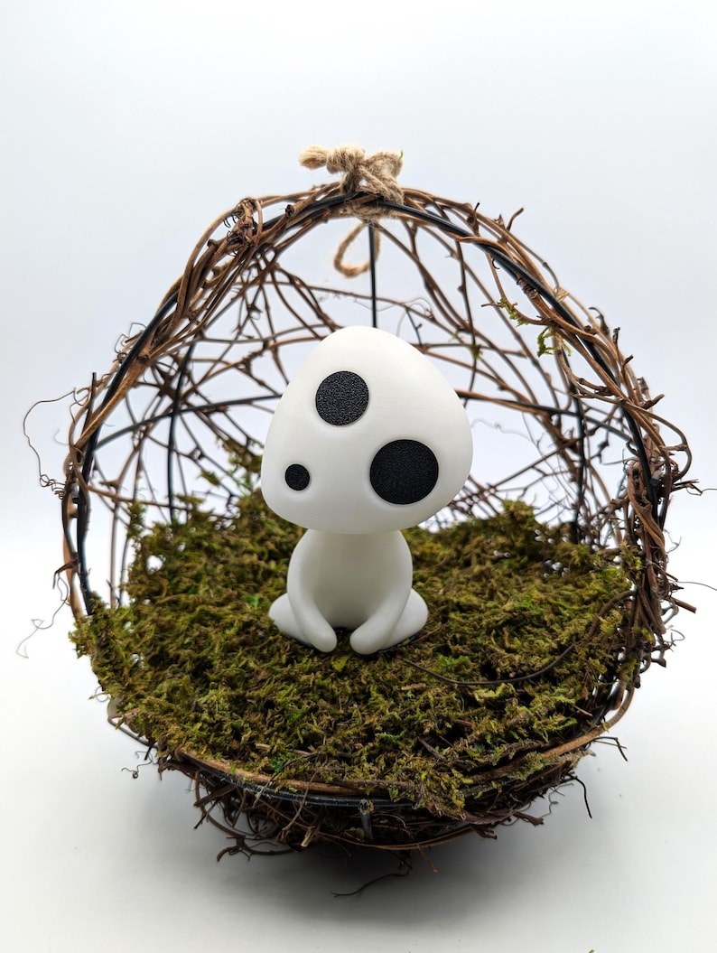 K&ouml;nnte beinhalten: Eine wei&szlig;e Figur mit drei schwarzen Punkten, die an einen Waldgeist erinnert, sitzt auf einem Bett aus gr&uuml;nem Moos in einer gewebten Zweigkugel. Die Kugel wird von einer nat&uuml;rlichen Schnurschlaufe gehalten, was eine skurrile, von der Natur inspirierte Darstellung ergibt.
