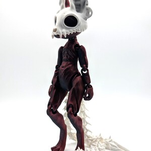 May include: A red and white articulated doll with a skeletal head and a long, segmented tail. The doll is standing on a white surface.