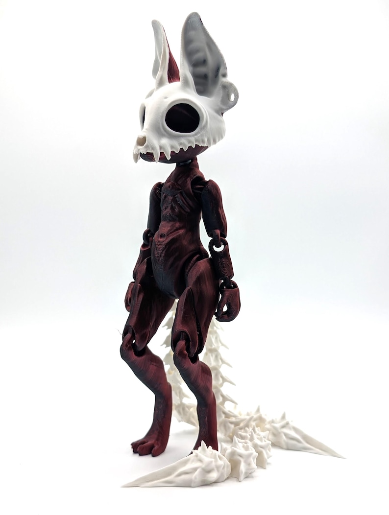 May include: A red and white articulated doll with a skeletal head and a long, segmented tail. The doll is standing on a white surface.