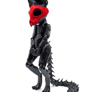 May include: Black articulated toy figure with a red skull mask. The figure has a long, spiky tail and is posed standing with its legs slightly bent.