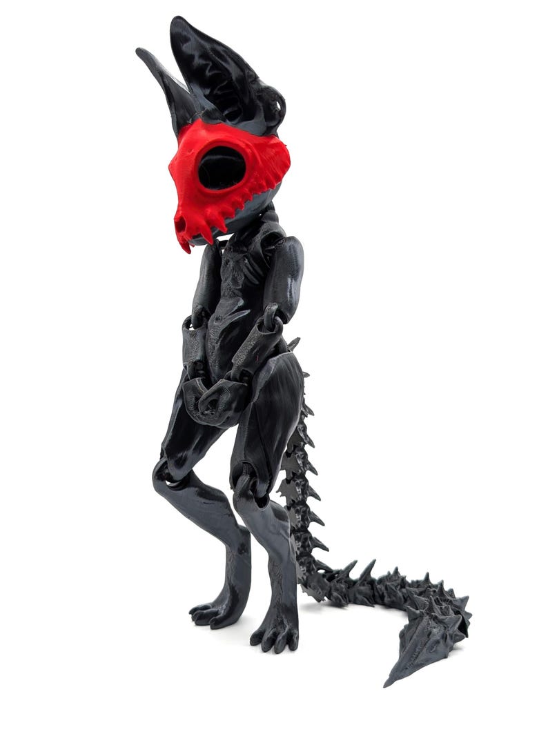 May include: Black articulated toy figure with a red skull mask. The figure has a long, spiky tail and is posed standing with its legs slightly bent.