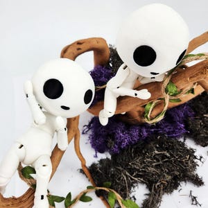 May include: Two white, articulated dolls with large black eyes, perched on a brown branch with green leaves. The dolls are surrounded by purple and black moss, creating a whimsical forest scene.