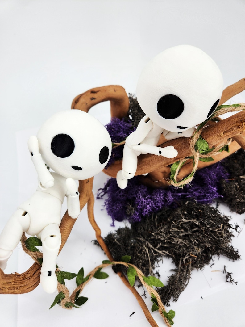 May include: Two white, articulated dolls with large black eyes, perched on a brown branch with green leaves. The dolls are surrounded by purple and black moss, creating a whimsical forest scene.