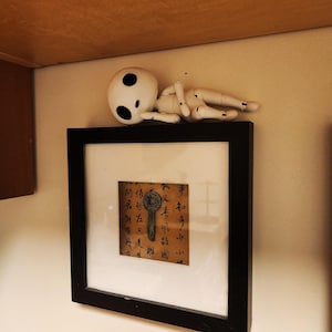 May include: A framed artwork with a black border, featuring a brown paper with calligraphy and a key illustration. A white, doll-like figure with black eye spots rests above the frame. The artwork is mounted on a beige wall.