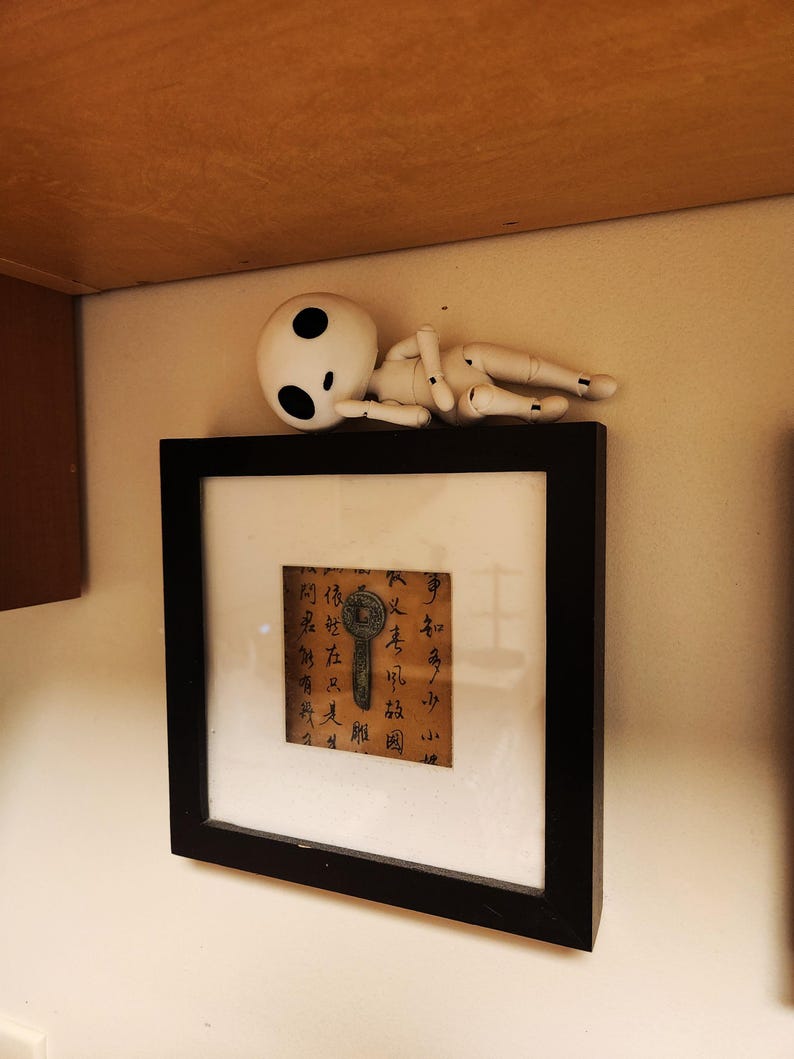 May include: A framed artwork with a black border, featuring a brown paper with calligraphy and a key illustration. A white, doll-like figure with black eye spots rests above the frame. The artwork is mounted on a beige wall.