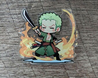 Op de afbeelding: Een heldere acryl bedel met een chibi-stijl karakter met groen haar en kleding, met een zwaard. Het karakter is omringd door oranje vlammen. Twee extra zwaarden zijn zichtbaar. De bedel ligt op een houten oppervlak.