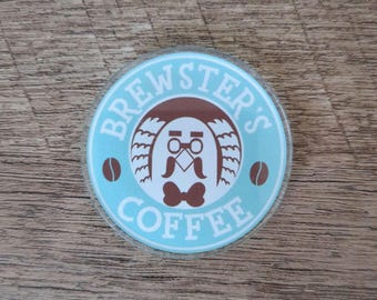 May include: A round coaster with a light blue background featuring the text "Brewster's Coffee" in white. The center design includes a brown cartoon character with a mustache and bow tie, and two coffee beans.