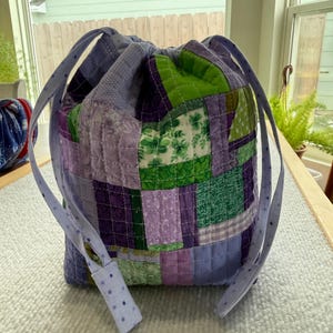 May include: A patchwork drawstring bag in shades of purple and green. The bag features a quilted design with various fabric squares sewn together. The drawstring and handle are a light purple with small blue polka dots. The bag is sitting on a white surface.