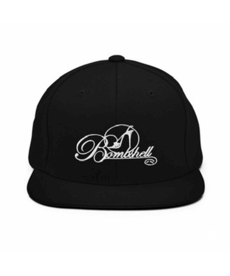 以下が含まれることがあります： 黒いベースボールキャップ、つばは平らです。「Bombshell」の文字が白で書かれており、ハイヒールの靴のグラフィックがデザインに組み込まれています。キャップは構造的なフィット感と丸みを帯びたクラウンを備えています。