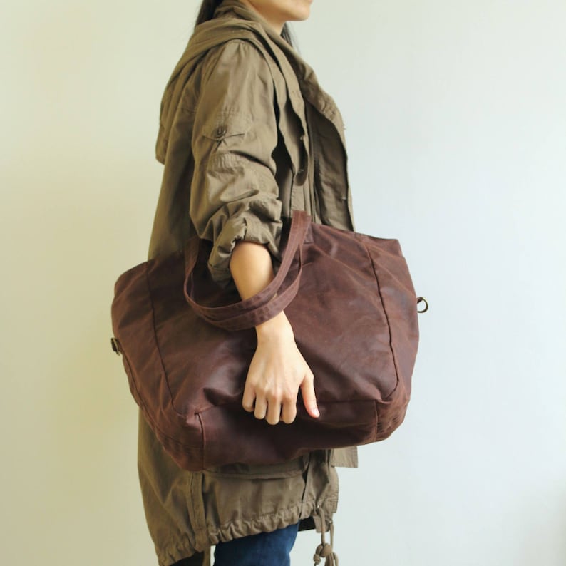 Puede incluir: Un bolso tote grande, marr&oacute;n, con dos asas. El bolso est&aacute; hecho de un material texturizado y tiene forma rectangular. El bolso lo lleva una persona que lleva una chaqueta verde.
