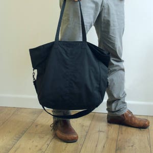 Puede incluir: Un bolso tote negro grande con asas largas y una correa cruzada desmontable. El bolso est&aacute; hecho de una tela duradera y tiene un dise&ntilde;o simple y funcional. El bolso se sostiene frente a una persona que lleva pantalones grises y zapatos marrones.