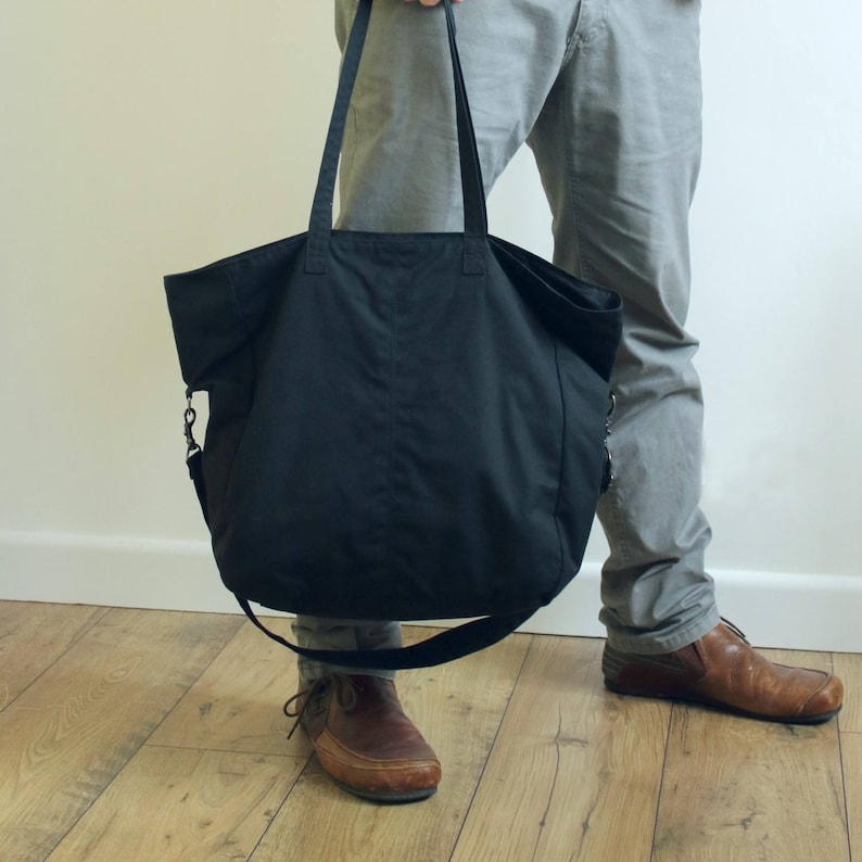 Puede incluir: Un bolso tote negro grande con asas largas y una correa cruzada desmontable. El bolso est&aacute; hecho de una tela duradera y tiene un dise&ntilde;o simple y funcional. El bolso se sostiene frente a una persona que lleva pantalones grises y zapatos marrones.