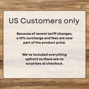 Puede incluir: Un letrero rectangular con borde de madera y fondo beige claro. El texto "US Customers only" est&aacute; en la parte superior. Debajo, el texto explica un recargo del 10% debido a los cambios arancelarios, sin sorpresas al finalizar la compra.