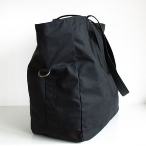 Puede incluir: Un bolso tote negro grande con base rectangular y parte superior redondeada. El bolso tiene correas largas y un anillo de metal plateado en el lateral. El bolso est&aacute; hecho de una tela resistente.