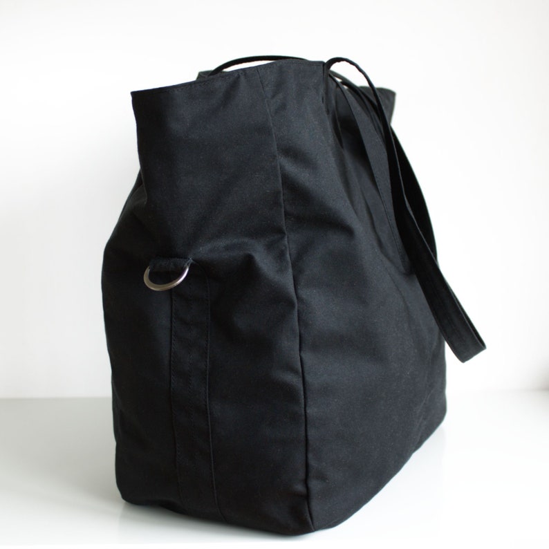 Puede incluir: Un bolso tote negro grande con base rectangular y parte superior redondeada. El bolso tiene correas largas y un anillo de metal plateado en el lateral. El bolso est&aacute; hecho de una tela resistente.