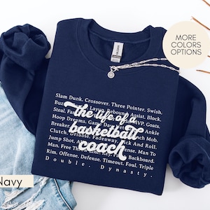 Könnte beinhalten: Marineblaues Sweatshirt mit weißem Text, der "the life of a basketball coach" und eine Liste von Basketballbegriffen zeigt. Eine silberne Halskette liegt über dem Sweatshirt. Die Worte "More Colors Options" stehen in einem Kreis rechts oben.