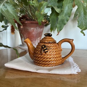 May include: A brown ceramic teapot with a beehive design and a bee applique. The teapot sits on a white cloth and has a curved handle and spout. A potted plant is in the background.
