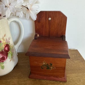 May include: A small, wooden wall-mounted salt box with a hinged lid and a decorative floral design. The box is stained in shades of brown and red, with a small hole at the top. The box is approximately 6 inches tall.