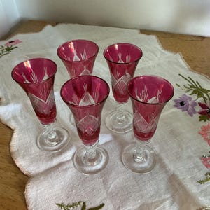 May include: A collection of five ruby red cordial glasses with clear, twisted stems. Each glass has a cut crystal design. The glasses are displayed on a white embroidered cloth.