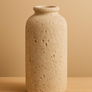 May include: A beige, textured vase with a cylindrical body and a narrow neck. The vase has a rough, porous surface and sits on a wooden shelf. The background is a solid beige colour. This decorative vase is suitable for home decor.