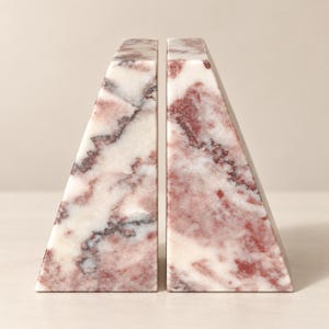 Puede incluir: Un par de sujetalibros triangulares hechos de mármol pulido, con un patrón de piedra natural que presenta vetas en tonos burdeos intenso, rosa suave y blanco crema a lo largo de las superficies lisas e inclinadas de cada pieza.