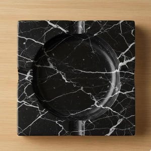 May include: Square black marble ashtray with white veining. The ashtray has a circular indentation in the center and a groove for resting cigarettes. The ashtray is made of a solid, heavy material.