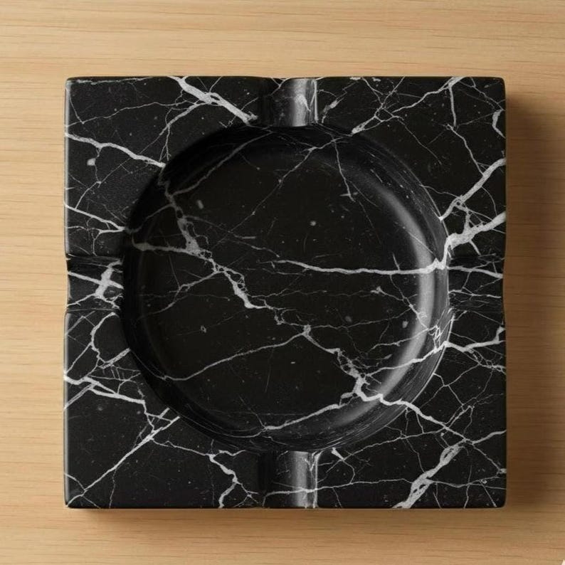 May include: Square black marble ashtray with white veining. The ashtray has a circular indentation in the center and a groove for resting cigarettes. The ashtray is made of a solid, heavy material.